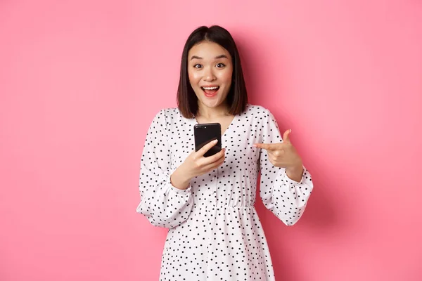Интернет-магазины и концепция красоты. Удивительная и счастливая азиатская женщина указывает на мобильный телефон, говорит об интернет-промо-предложение или приложение, стоя на розовом фоне — стоковое фото