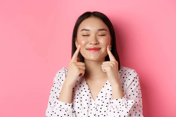 Beauty und Lifestylekonzept. Nahaufnahme einer schönen asiatischen Frau, die mit geschlossenen Augen über Wangen stochert, zufrieden lächelt und vor rosa Hintergrund steht — Stockfoto