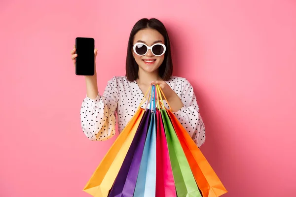 Attraktive Asiatin zeigt Smartphone-App und Einkaufstaschen, kauft online per App, steht vor rosa Hintergrund — Stockfoto