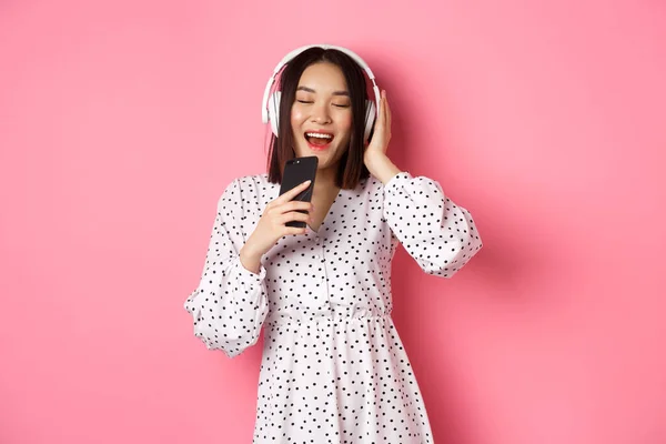 Mujer asiática linda jugando aplicación de karaoke, cantando en el teléfono móvil y el uso de auriculares, de pie en el vestido sobre fondo rosa — Foto de Stock