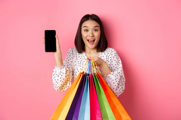 스마트폰 앱과 쇼핑백을 보여 주고, 앱을 통해 온라인 구매를 하고, 핑크 색 배경 위에 서 있는 매력적 인 아시아 여성 — 스톡 사진