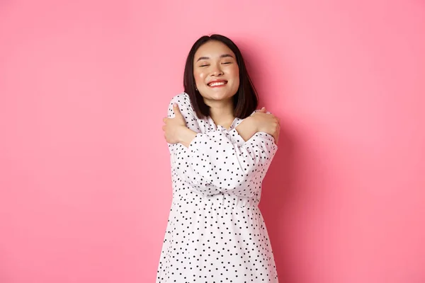 Vacker asiatisk kvinna som känner sig romantisk, kramar sig själv, blundar och ler med drömmande ansikte, står lycklig över rosa bakgrund — Stockfoto