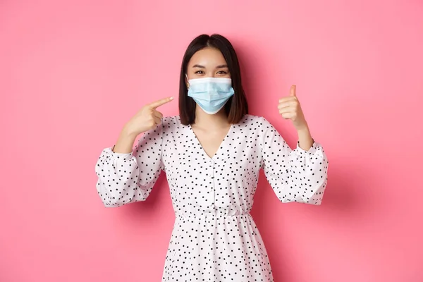 Coronavírus, distanciamento social e conceito de estilo de vida. Mulher asiática bonito apontando para máscara facial, mostrando polegar para cima, apoio usando medidas preventivas de covid-19, fundo rosa — Fotografia de Stock