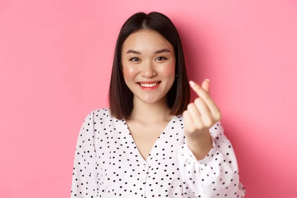 Conceito de beleza e estilo de vida. Close-up de linda mulher asiática mostrando sinal de coração, sorrindo e se sentindo romântico no dia dos namorados, de pé sobre fundo rosa — Fotografia de Stock