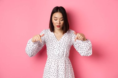 Romantik Asyalı kadın dikizliyor, parmakları dipte, ürün indirimine bakıyor, pembe arka planda duruyor.