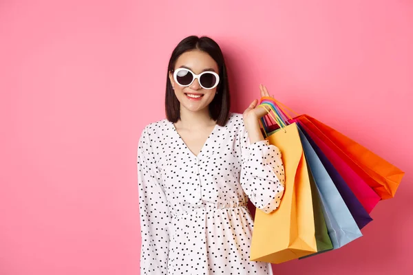 Mladá Asiatka v slunečních brýlích jde nakupovat, drží tašky z obchodů a obchodů a usmívá se, stojí nad růžovým pozadím — Stock fotografie