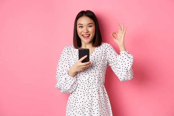 Online shopping och skönhet koncept. Nöjd asiatisk kvinnlig kund visar okej, gör inköp på internet på smartphone, står över rosa bakgrund — Stockfoto