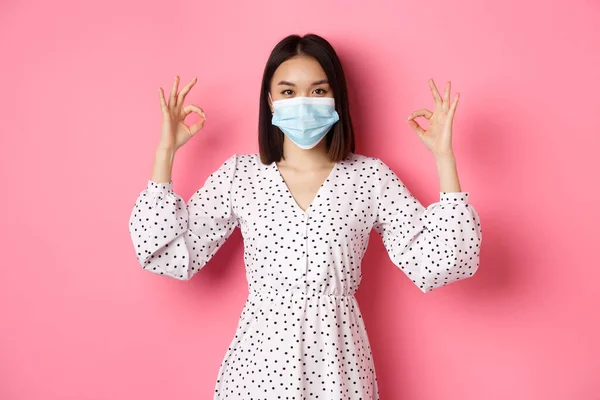 Coronavirus, sosial jarak dan konsep gaya hidup. Wanita asia yang puas bertopeng wajah menunjukkan tanda-tanda yang baik, menyetujui dan setuju, memuji pilihan yang baik, latar belakang merah muda — Stok Foto
