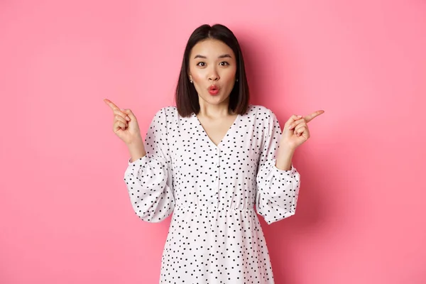 Hermosa mujer asiática haciendo elección en las compras, señalando los dedos hacia los lados y mostrando variantes, sonriendo a la cámara, de pie sobre el fondo rosa —  Fotos de Stock