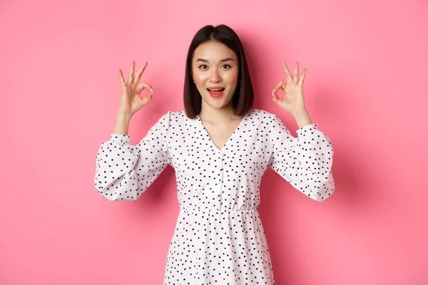 Jolie jeune femme asiatique en robe montrant le signe correct, louant et montrant l'approbation, regardant satisfait, debout sur fond rose — Photo