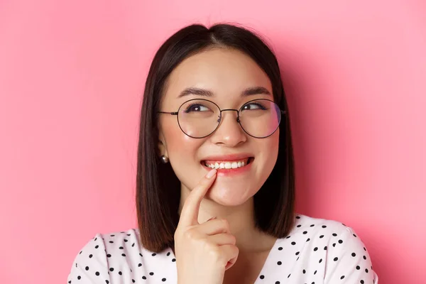 Headshot de femme asiatique mignonne dans des lunettes à la mode souriant, ayant une idée, pensant et regardant coin supérieur gauche, fond rose — Photo