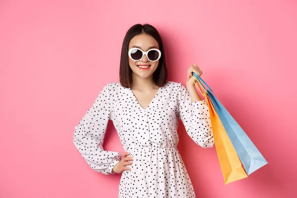 Attraktive lächelnde asiatische Frau mit Einkaufstaschen, Sonnenbrille und niedlichem Kleid vor rosa Hintergrund — Stockfoto