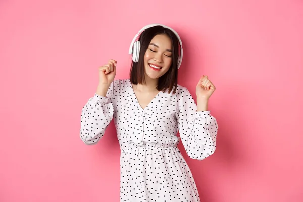 Feliz joven mujer asiática bailando y divirtiéndose, escuchando música en auriculares, de pie sobre fondo rosa — Foto de Stock