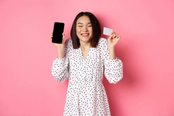 Радостная корейская женщина, показывающая экран смартфона и кредитную карту, платящая за интернет-заказ, демонстрирующая приложение для покупок онлайн, стоящая на розовом фоне — стоковое фото