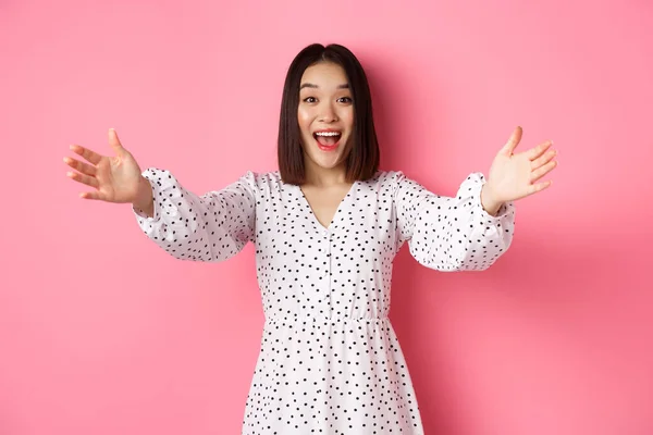 Boldog ázsiai lány nyújtja kezét a kamera, nyúl az ölelés, mosolygós és akar tartani valamit, áll át rózsaszín háttér ruhában — Stock Fotó