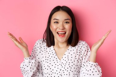 Şaşırmış ve mutlu Asyalı kadın sevinçten havalara uçuyor, ellerini yana açıyor ve gülümsüyor, harika haberler duyuyor, pembe arka planda duruyor.