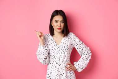 Hayal kırıklığına uğramış Asyalı kız arkadaş şikayet ediyor, kameraya bakıyor ve mızmızlanıyor, sol üst köşedeki fotokopi alanını işaret ediyor, pembe arka plan üzerinde duruyor.