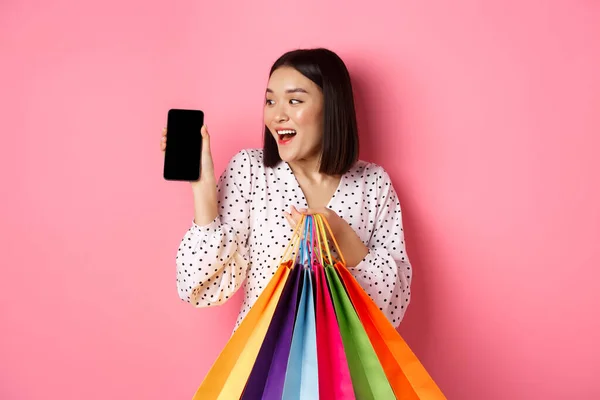 스마트폰 앱과 쇼핑백을 보여 주고, 앱을 통해 온라인 구매를 하고, 핑크 색 배경 위에 서 있는 매력적 인 아시아 여성 — 스톡 사진
