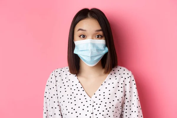 Covid-19, pandemi och livsstilskoncept. Förvånad asiatisk kvinna i ansiktsmask höja ögonbryn, stirrar på kameran förvånad, står mot rosa bakgrund — Stockfoto