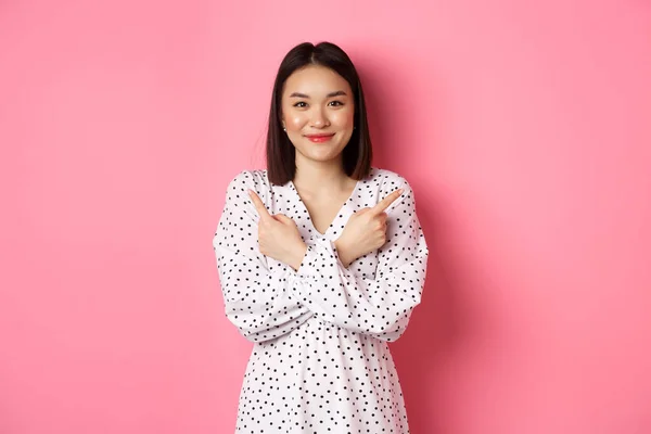 Belle fille asiatique en robe romantique pointant les doigts sur le côté, montrant deux variantes sur le shopping, souriant à la caméra, debout sur fond rose — Photo