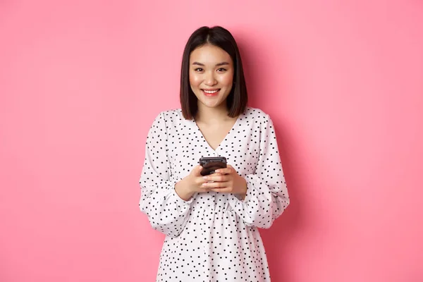 Piękna azjatycka pani czyta wiadomości i uśmiecha się, za pomocą telefonu komórkowego, stojąc w uroczej sukience na różowym tle — Zdjęcie stockowe
