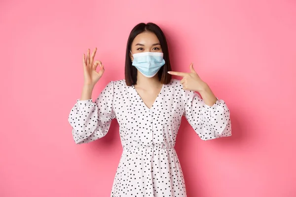 Coronavirus, social distancing and lifestyle concept. Cute asian woman pointing at face mask, showing okay sign, support using preventive measures from covid-19, pink background — Stock Photo, Image