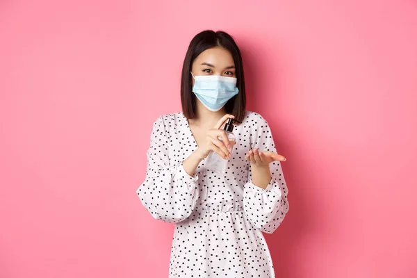 Covid-19, világjárvány és életmód koncepció. Aranyos ázsiai nő tiszta kezek fertőtlenítő, használ fertőtlenítő és visel orvosi maszk, felett áll rózsaszín háttér — Stock Fotó