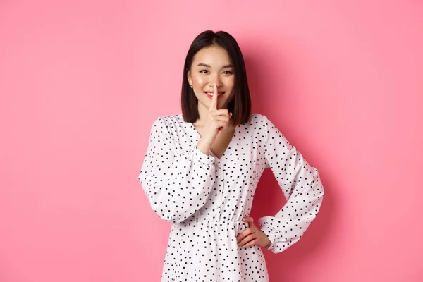 Hermosa mujer coreana en vestido de moda pidiendo mantener el secreto, silenciando con una sonrisa suave, planeando una sorpresa, de pie sobre el fondo rosa —  Fotos de Stock