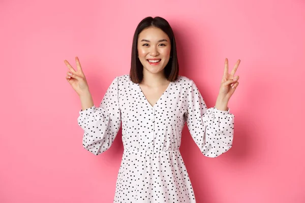 옷을 입고 미소짓고 있는 귀여운 아시아인 브 라무네 소녀, 카와 비의 평화 표시를 보여 주며 핑크 색 배경 위에 서 있는 행복 해 보이는 모습 — 스톡 사진