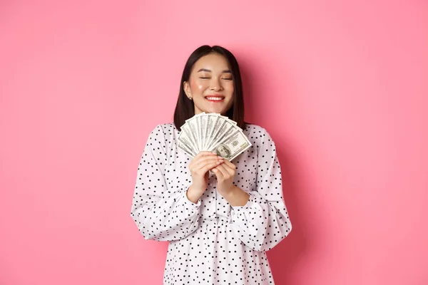 Концепция покупок. Счастливая и довольная азиатка выигрывает призовые деньги, показывая доллары и ликование, стоя на розовом фоне — стоковое фото