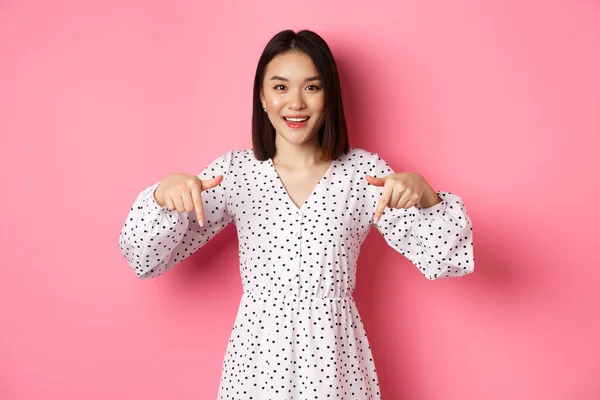 Olha para ali. bonito ásia mulher no vestido apontando dedos para baixo no cópia espaço mostrando produto desconto e sorrindo de pé sobre rosa fundo — Fotografia de Stock