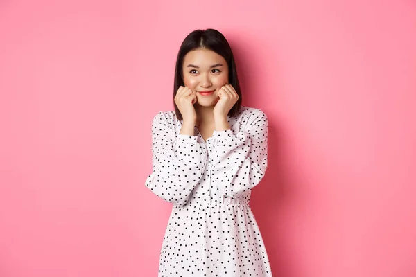 Χαριτωμένο και ντροπαλό κορίτσι από την Ασία κοκκίνισμα, αγγίζοντας τα μάγουλα και κοιτάζοντας αριστερά στο χώρο αντίγραφο ανόητο, στέκεται κατά ροζ φόντο — Φωτογραφία Αρχείου
