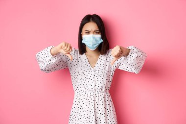 Coronavirus, sosyal uzaklık ve yaşam tarzı konsepti. Yüz maskesi takmış hayal kırıklığına uğramış Asyalı kadın yargılayıcı görünüyor, baş parmaklarını göstermiyor ve katılmıyor, pembe arka planda duruyor.