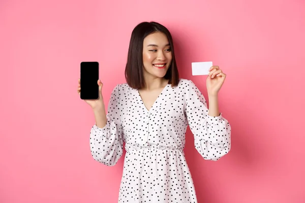 Szczęśliwy młody Azji kobieta internet zakupy, pokazując ekran smartfona i patrząc zadowolony na karty kredytowej, stojąc nad różowym tle — Zdjęcie stockowe