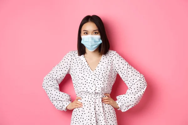 Covid-19, concept de quarantaine et style de vie. Mignonne asiatique femme portant un masque médical et robe, se protéger contre le coronavirus, debout sur fond rose — Photo