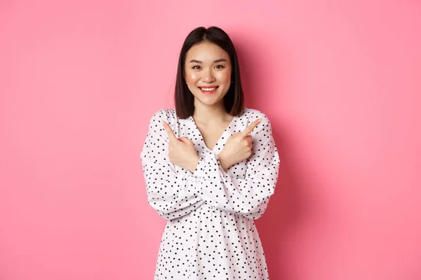Menina asiática bonita em vestido romântico apontando os dedos lateralmente, mostrando duas variantes em compras, sorrindo para a câmera, de pé contra o fundo rosa — Fotografia de Stock