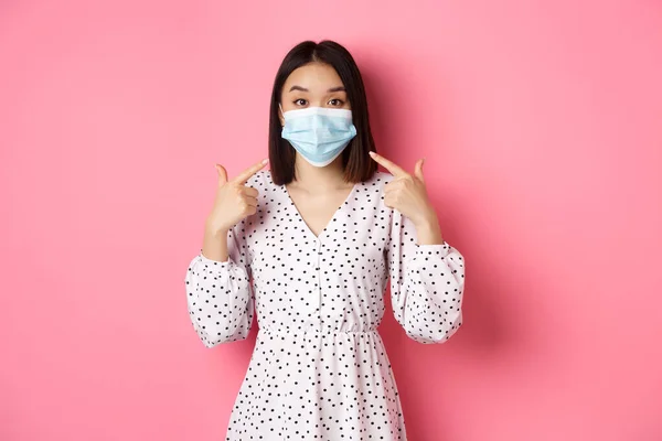 Coronavirus, έννοια της κοινωνικής απόστασης και του τρόπου ζωής. Χαριτωμένη Ασιάτισσα που δείχνει τη μάσκα προσώπου, ζητώντας της να χρησιμοποιήσει μέτρα εναντίον του covid-19, στέκεται πάνω από ροζ φόντο — Φωτογραφία Αρχείου