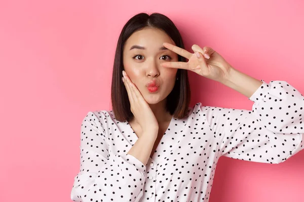 Concepto de belleza y estilo de vida. primer plano de bastante asiático mujer mostrando paz signo y tocando mejilla, sonriendo feliz a cámara, de pie sobre rosa fondo —  Fotos de Stock