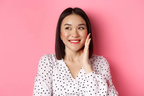 얼굴을 붉히고, 뺨을 만지고, 왼쪽을 보고, 분홍빛 배경 위에 서서 웃고 있는 아시아 여자 — 스톡 사진