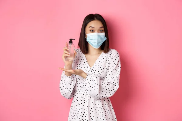Covid-19, salgın ve yaşam tarzı konsepti. Elbise ve tıbbi maske takan güzel Koreli kadın el dezenfektanı gösteriyor, antiseptik öneriyor, pembe arka planda duruyor. — Stok fotoğraf
