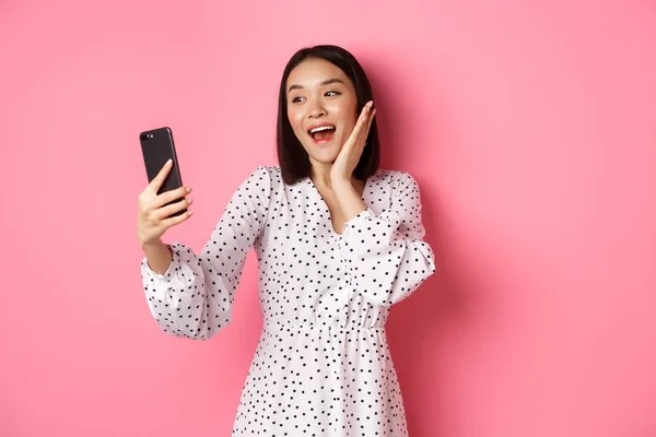 Piękna azjatycka dziewczyna za pomocą aplikacji filtrów fotograficznych i biorąc selfie na smartfonie, pozowanie w cute sukienka na różowym tle — Zdjęcie stockowe