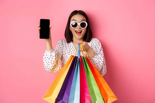 Atraktivní asijské žena ukazuje smartphone app a nákupní tašky, nákup on-line prostřednictvím aplikace, stojící nad růžovým pozadím — Stock fotografie