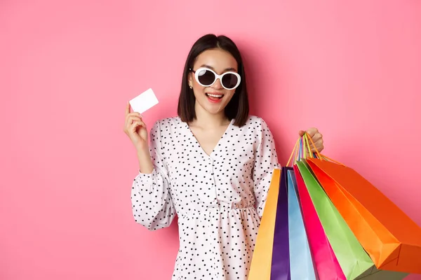 Krásná asijská žena v slunečních brýlích jít nakupovat, držení tašky a ukazuje kreditní kartu, stojící nad růžovým pozadím — Stock fotografie