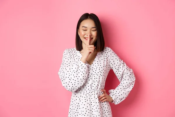 Hermosa mujer coreana en vestido de moda pidiendo mantener el secreto, silenciando con una sonrisa suave y ojos cerrados, de pie sobre el fondo rosa —  Fotos de Stock