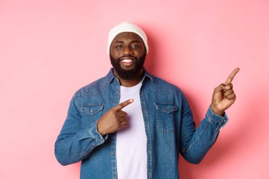 Mutlu Afro-Amerikalı adam gülümsüyor, parmaklarını doğru gösteriyor ve tanıtım yapıyor, duyuru yapıyor, pembe arka planda duruyor.