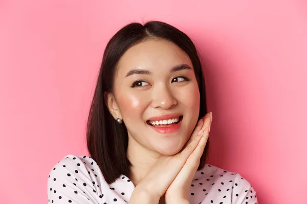 Concept beauté et soin de la peau. Headshot de femme asiatique adorable et rêveuse regardant à gauche, souriant et imagerie, debout sur fond rose — Photo