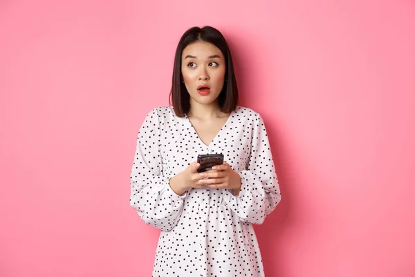 网上购物。困惑的亚洲女孩拿着智能手机，呆呆地看着左上角，站在粉色的背景上 — 图库照片