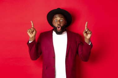 Kış tatili ve alışveriş konsepti. Etkilenmiş Afro-Amerikan adam parti kıyafeti içinde parmaklarını havaya kaldırıyor, Noel tanıtımına bakıyor, kırmızı arka planda duruyor.