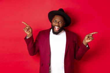 Kış tatili ve alışveriş konsepti. Neşeli siyahi adam gülümsüyor ve iki reklam gösteriyor, fotokopi alanlarını işaret ediyor, kırmızı arka plan