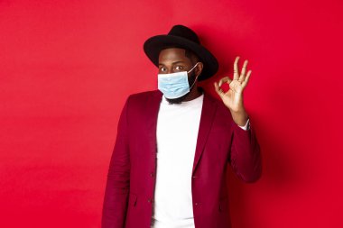 Covid 19 ve moda konsepti. Şapkalı ve ceketli şık Afro-Amerikan adam, yüz maskesi takıyor ve kırmızı arka planda duruyor.